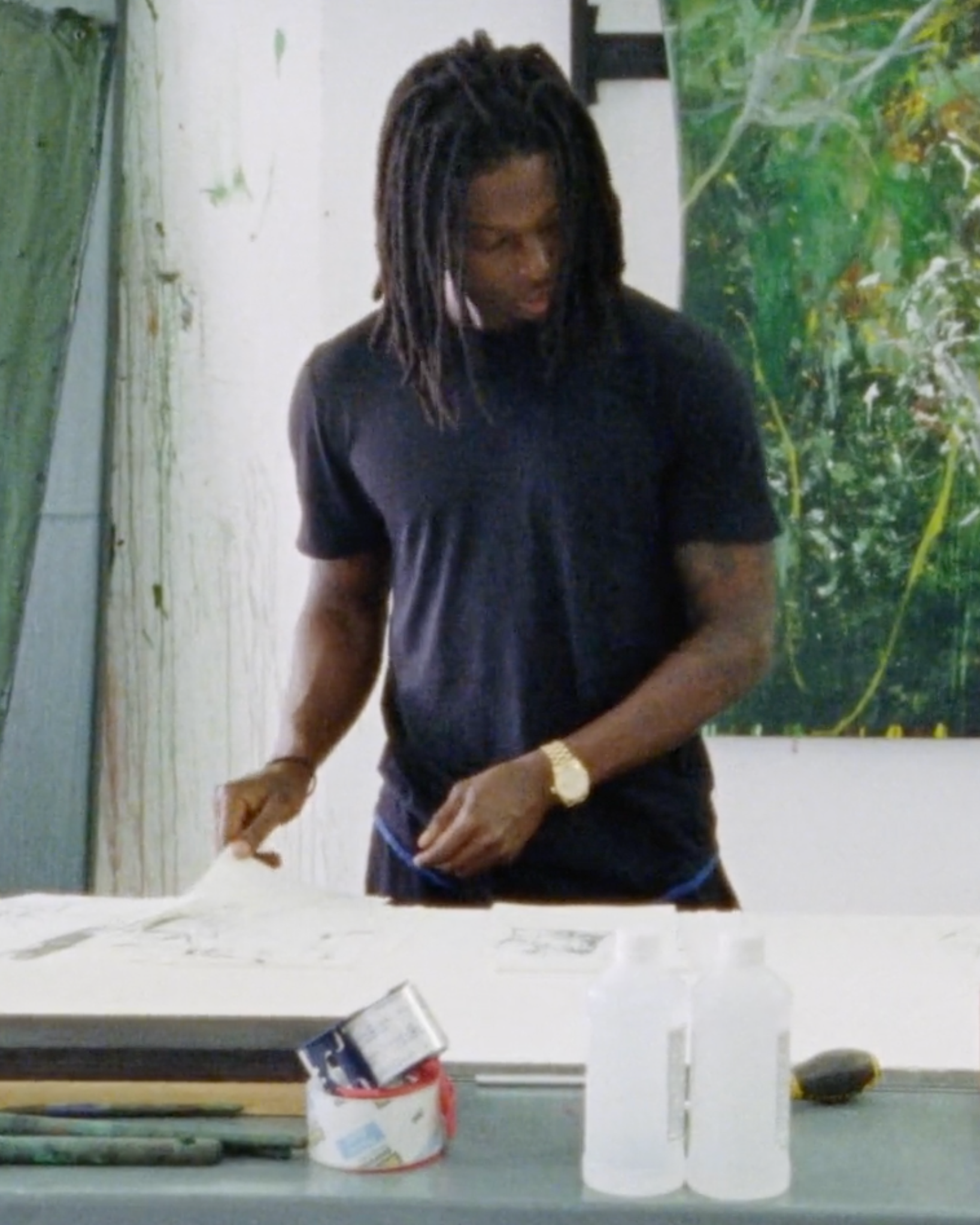 A person with long dreadlocks wearing a black t-shirt stands at a table, looking down at artwork or sketches. Art supplies and bottles are on the table, and a large painting with green tones is in the background.