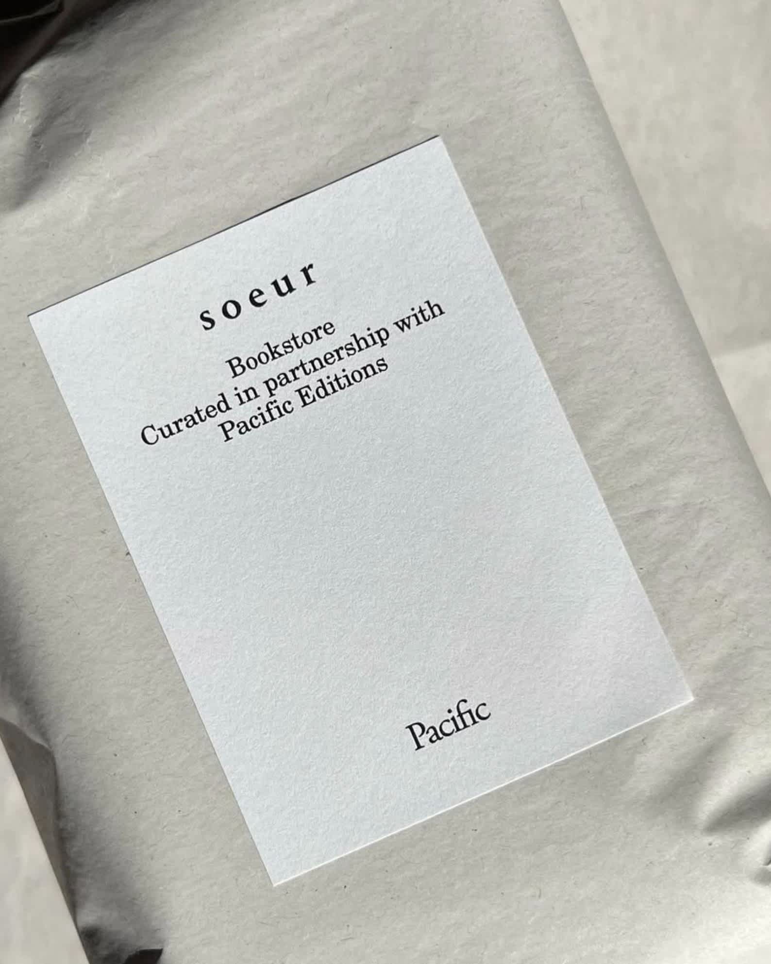 A close-up of a white label on a gray package reads: soeur Bookstore Curated in partnership with Pacific Editions Pacific. The background is softly blurred.