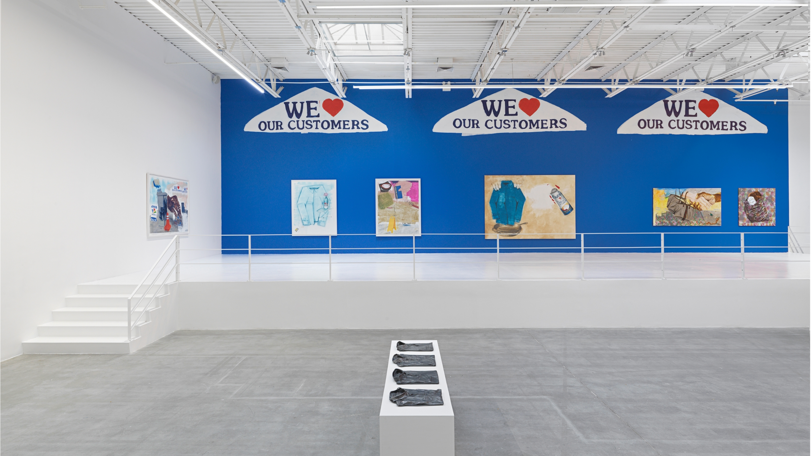 Installation shot from Pat Phillips' show at Jeffrey Deitch titled, "Consumer Reports". The room is mostly empty except for a white pedestal in the center which displays four folded shirts. A set of stairs on the left of the room leads up to a railed platform. The wall behind the platform is painted bright blue and has five paintings hung beneath three large-scale murals of dry-cleaning hangers.