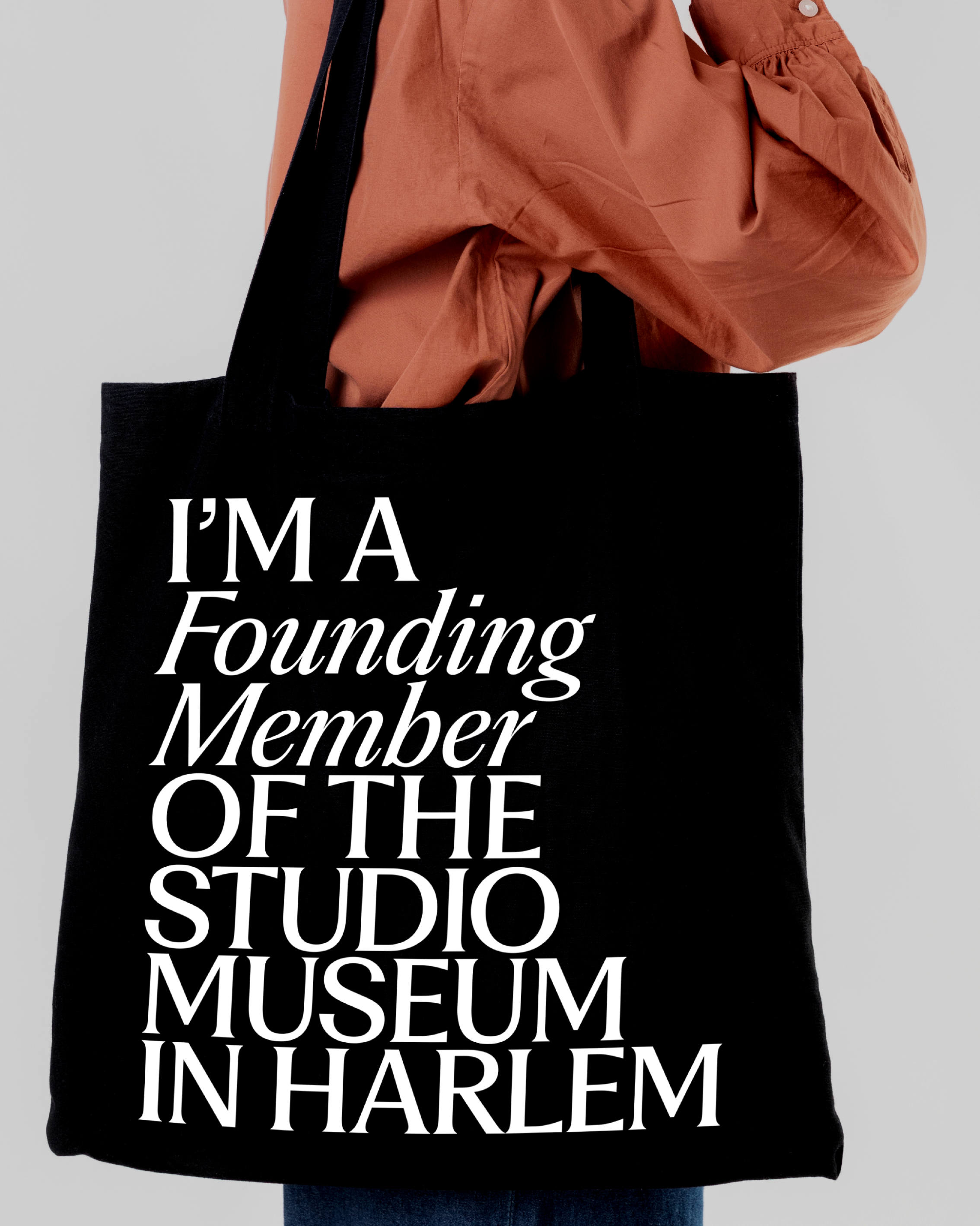 A person carries a black tote bag with bold white text that reads, IM A Founding Member OF THE STUDIO MUSEUM IN HARLEM.