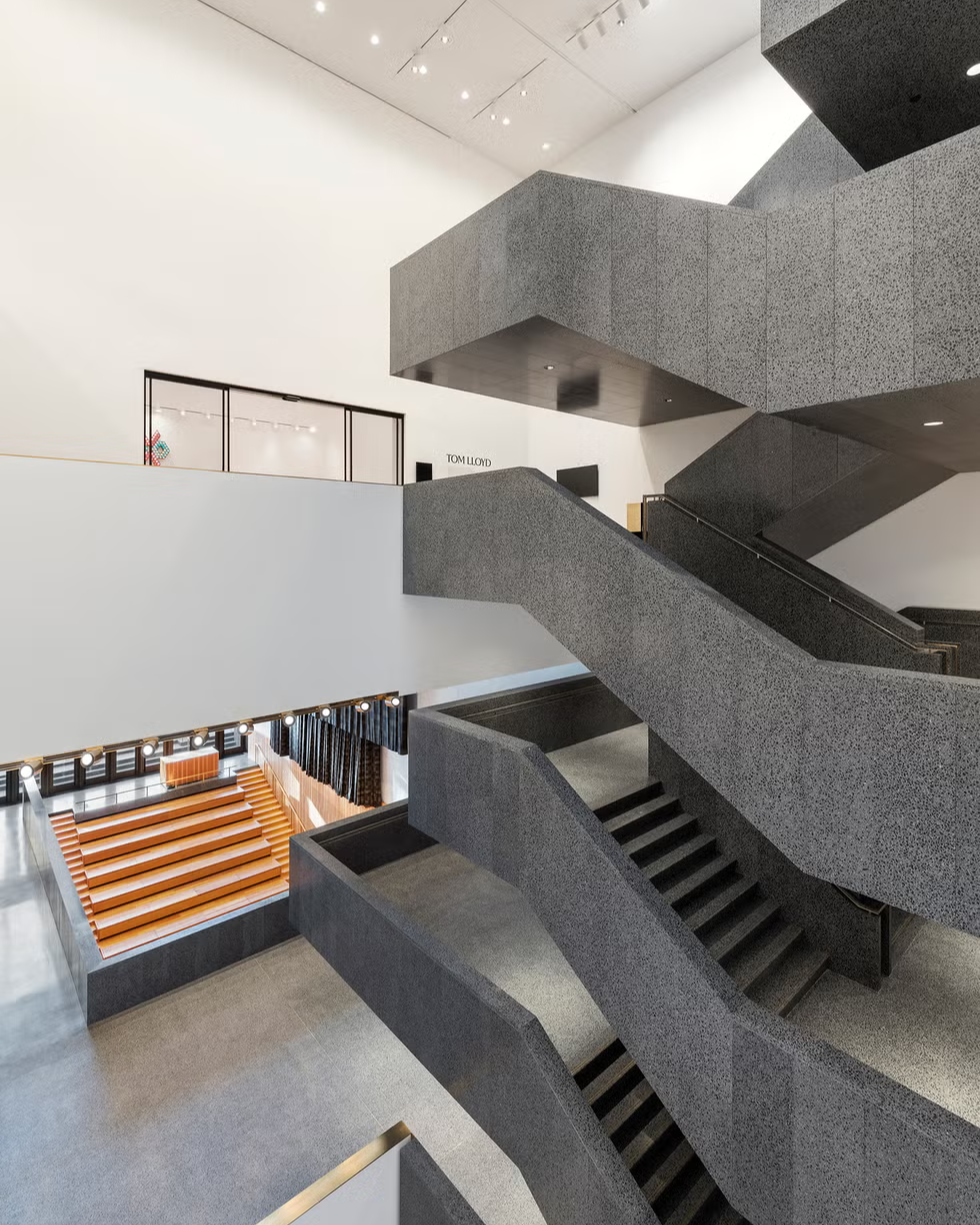 A modern interior with multiple angular, gray staircases crossing each other, white walls, and an open seating area with wooden benches on the lower level. Large windows and high ceilings create a spacious atmosphere.