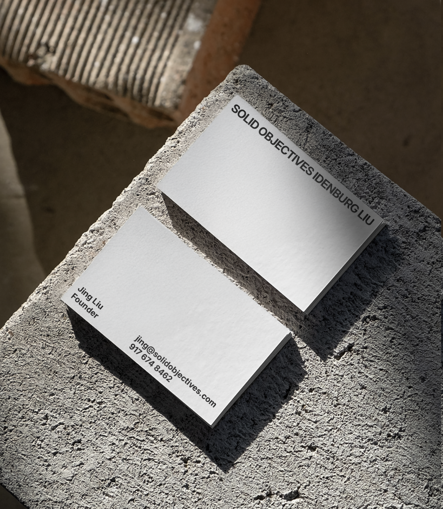 Two minimalist white business cards with black text rest on a textured concrete surface. One card displays the company name SOLID OBJECTIVES | DENBURG LIU, while the other shows contact details for Jing Liu, Founder.
