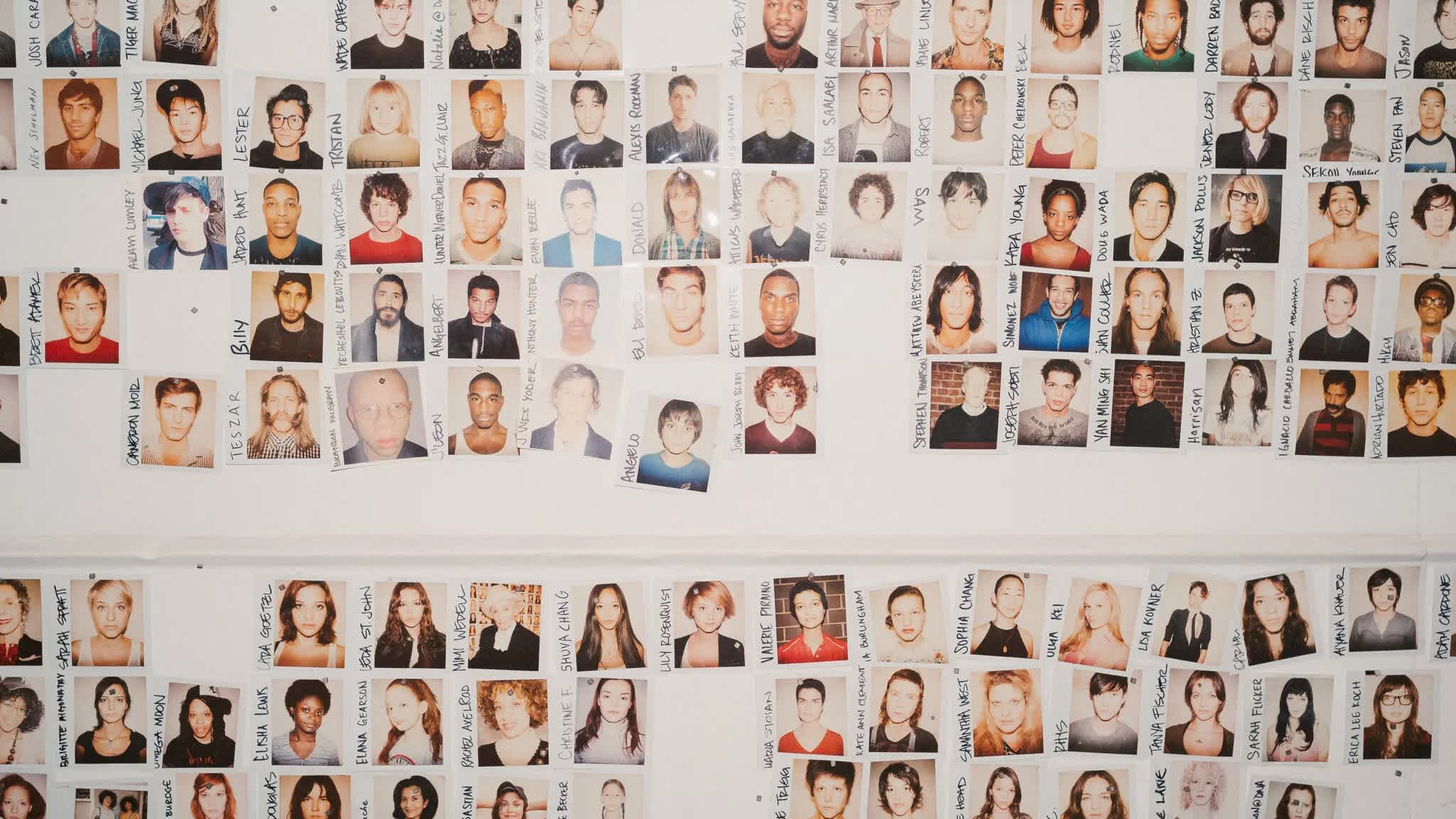 Photograph of a casting wall. The wall is covered in various polaroids of people from the chest up. There is an overall warm cast to the image.