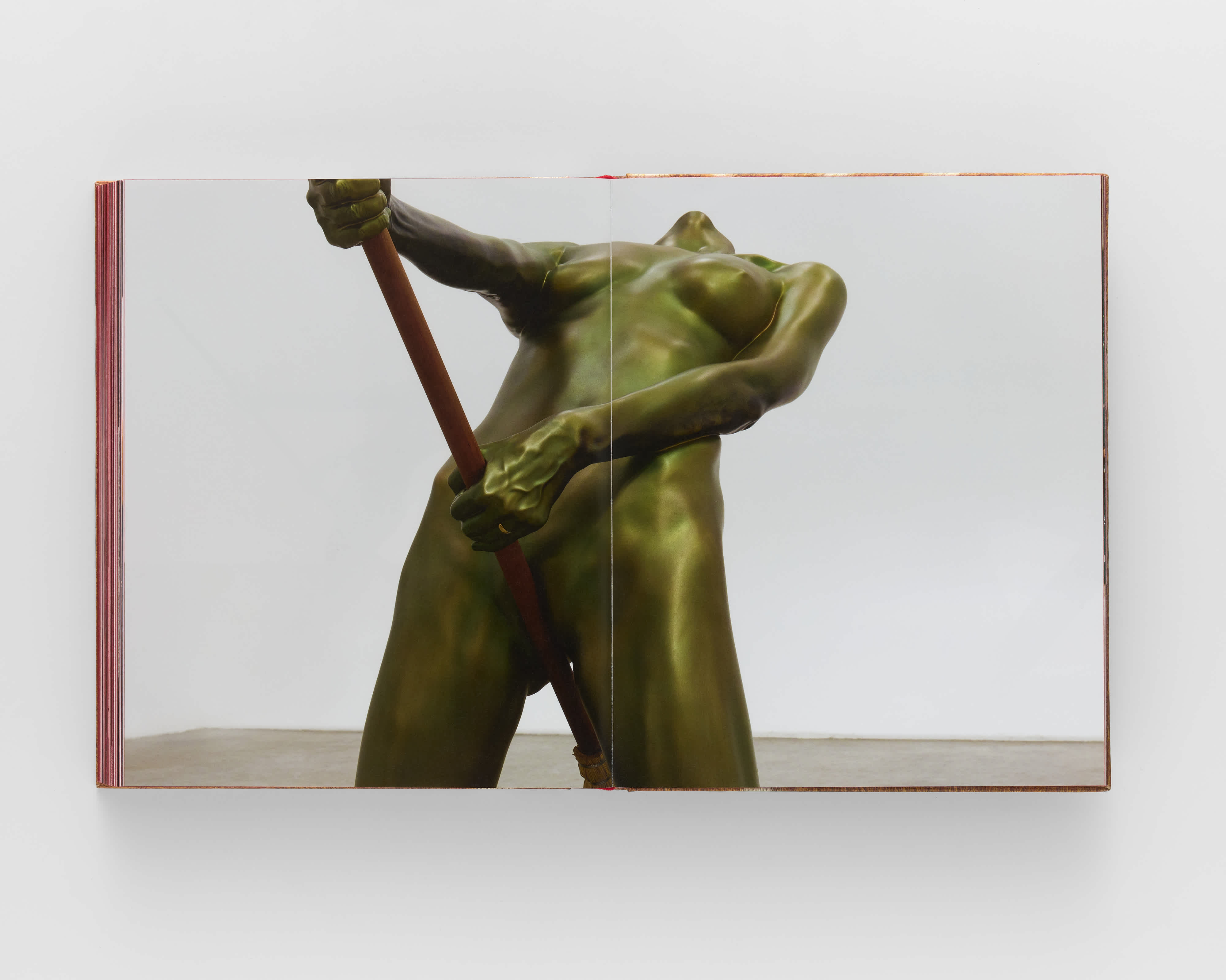 An open book on a grey background. A full-bleed, centerfold image of a sculpture takes up both pages. The sculpture is a greenish-metal casting of the human form riding a broom.