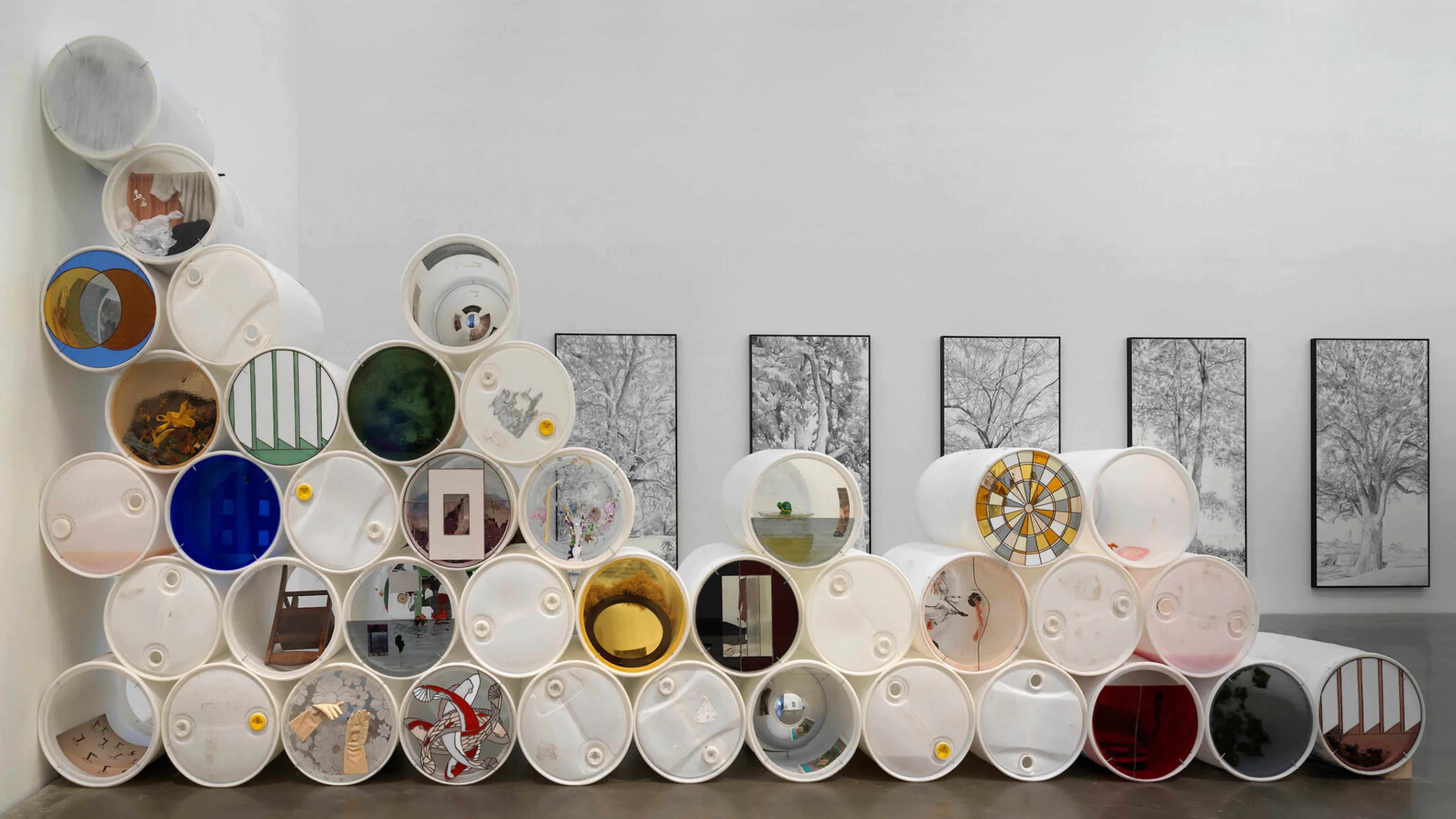 Installation shot from an exhibition at The New Museum. White rain barrels with various artworks replacing the lids, stacked against a wall. In the background there are five black and white images of trees hung on the far wall.
