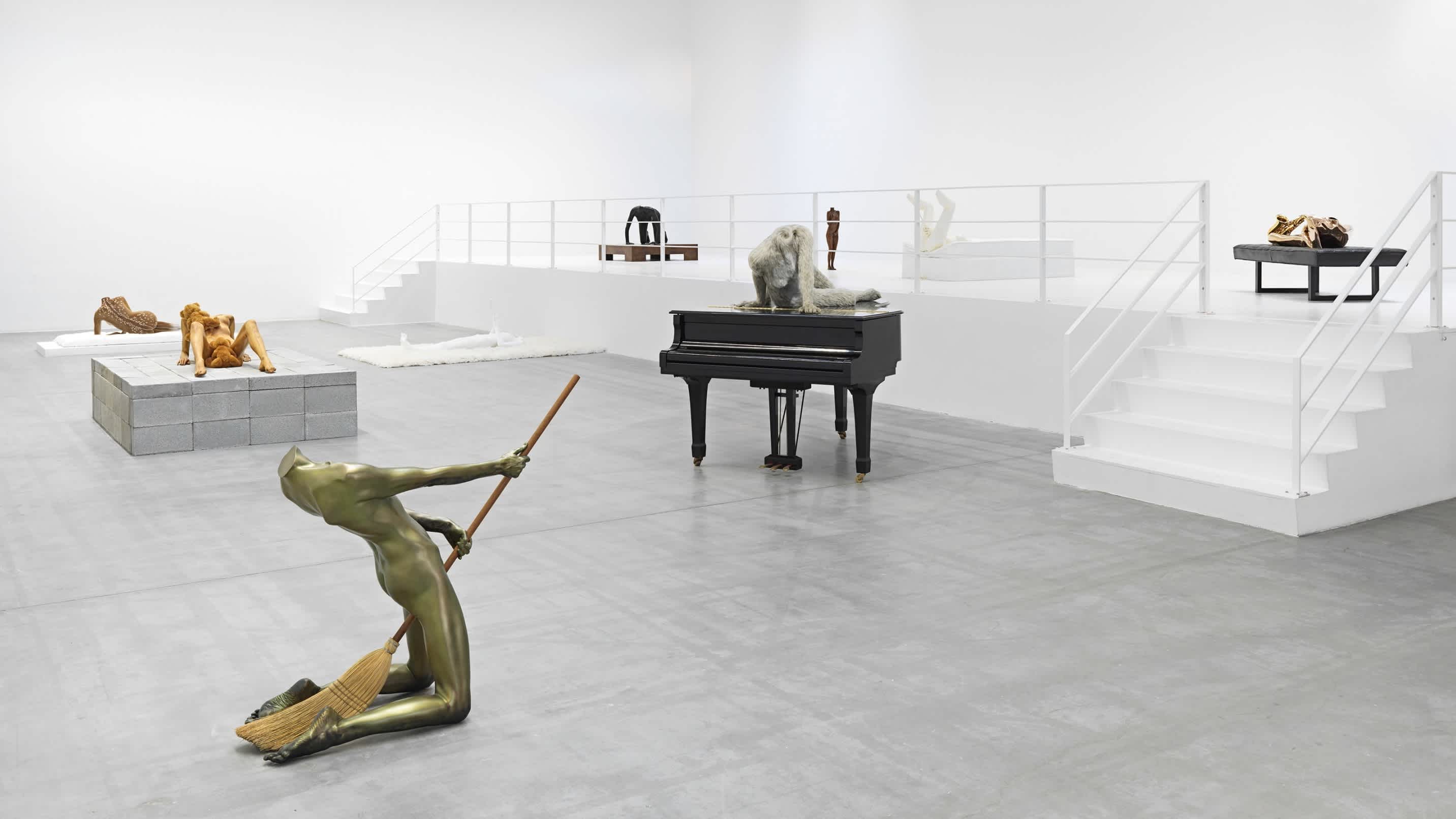 Eight sculptures of the manipulated human body are positioned throughout a concrete-floored, and white-walled room. 
