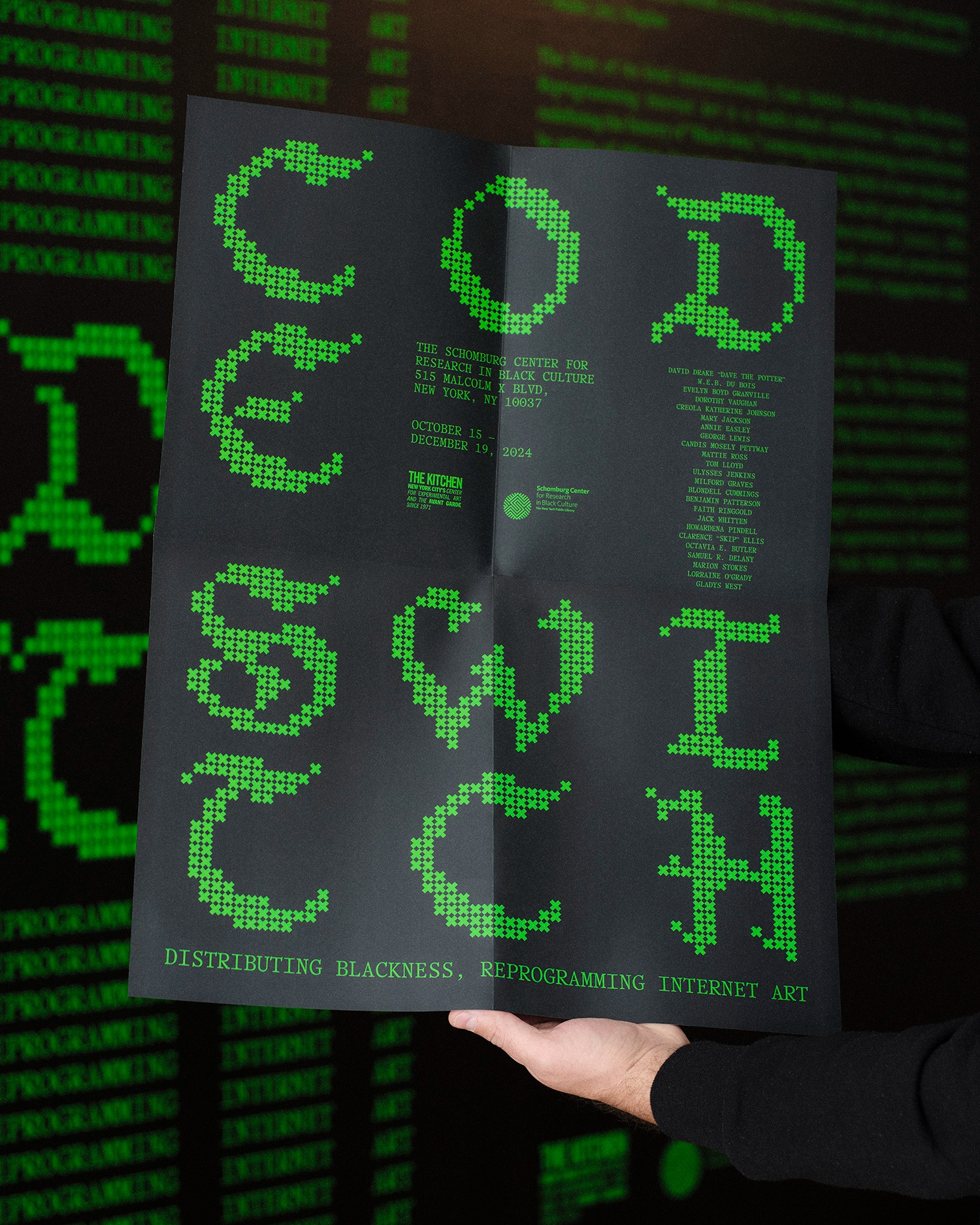 A person holds a black poster with large, pixelated green letters and text that reads “CODE SWITCH: Distributing Blackness, Reprogramming Internet Art.” A digital green pattern is visible in the background.