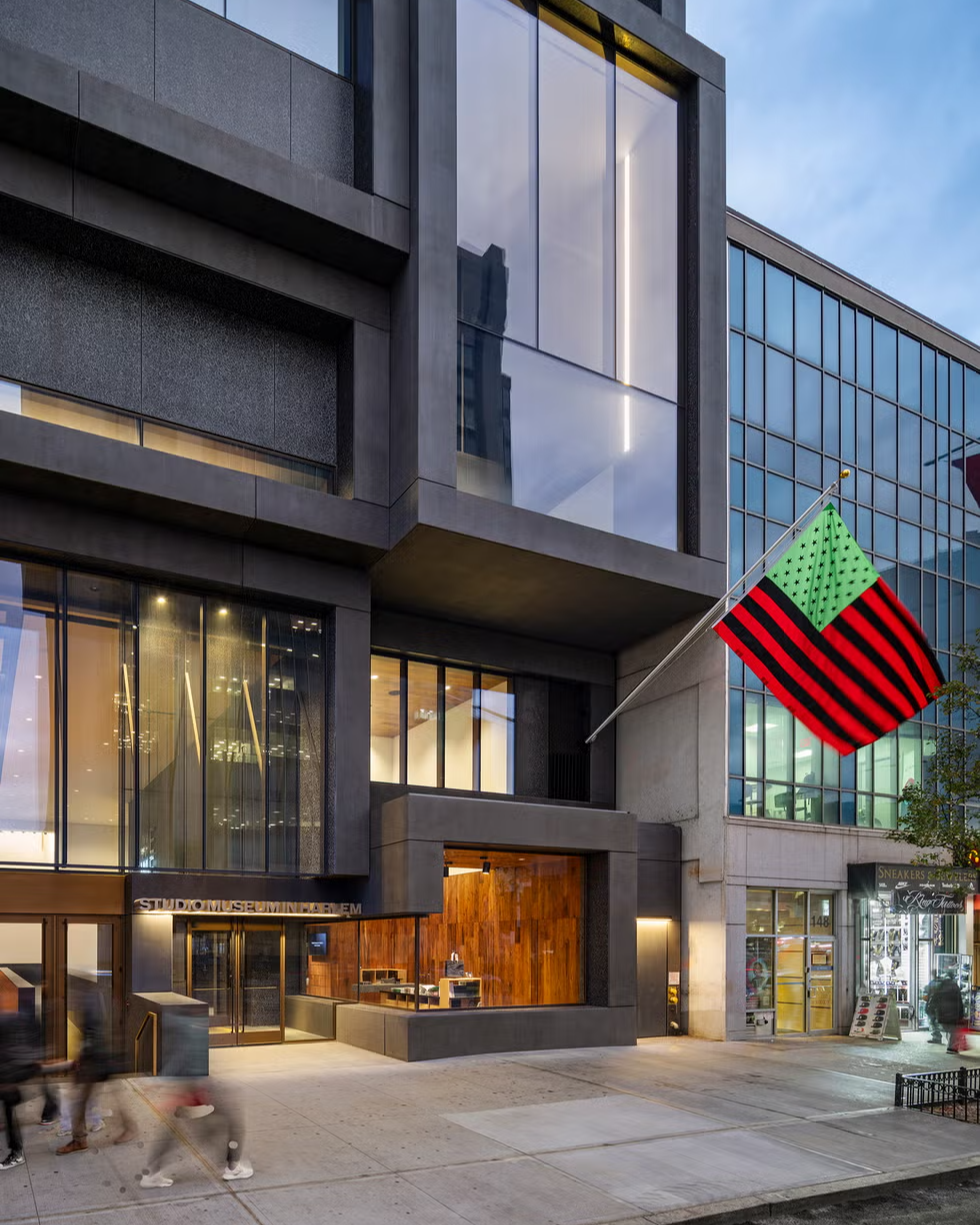 A modern building with large glass windows and concrete features stands on a city street. A red, black, and green flag with stars hangs outside, and blurred pedestrians walk on the sidewalk in front.