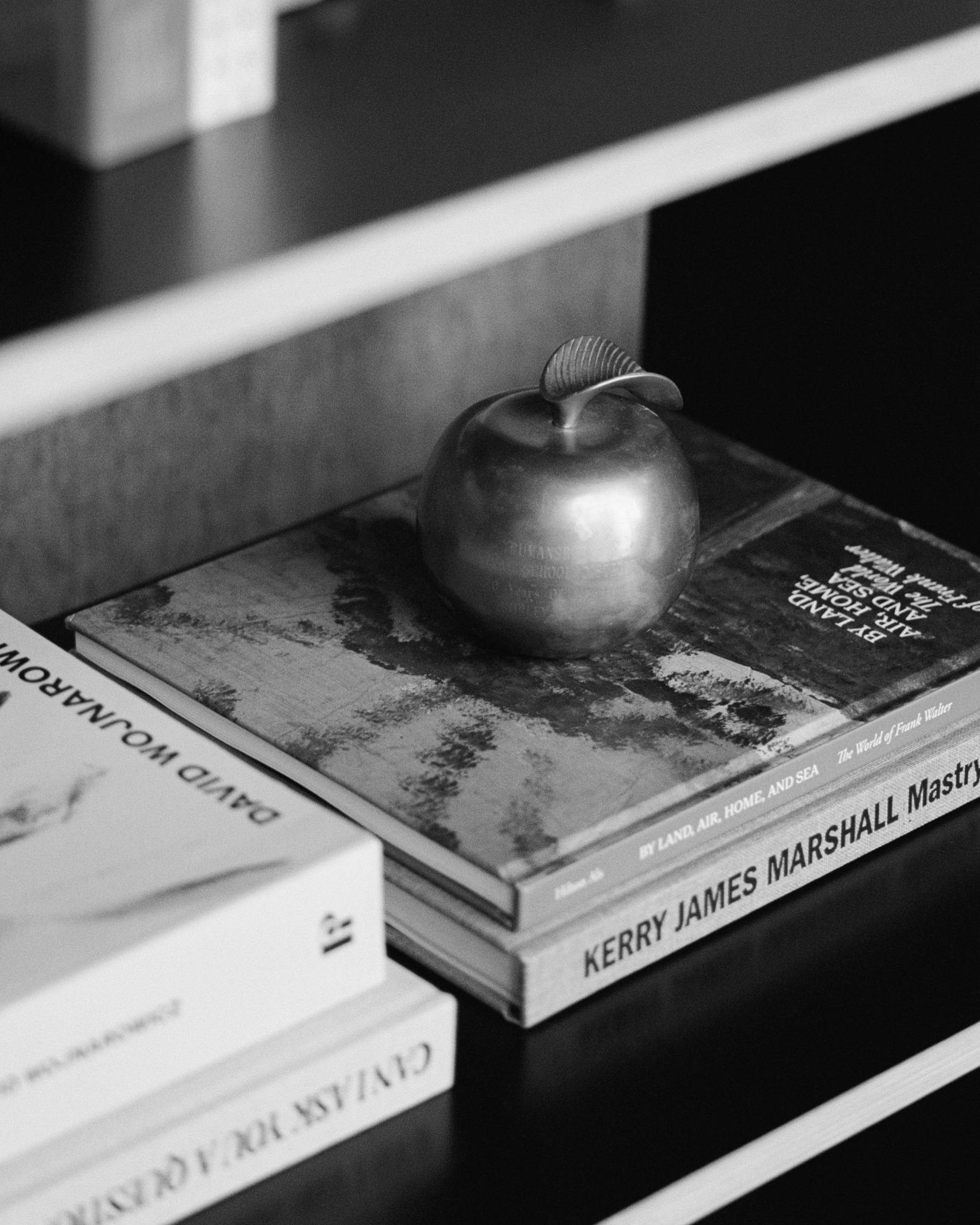 A metallic apple ornament sits on top of a stack of art books, including titles by Kerry James Marshall and Olivo Barbieri, on a dark shelf. The image is in black and white.