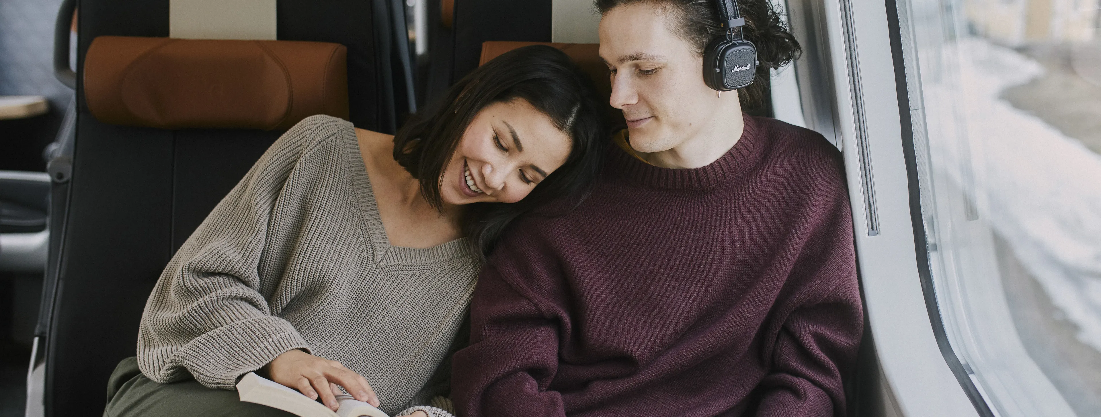 Pariskunta vierekkäin junassa. Nainen lukee kirjaa ja mies kuuntelee musiikkia kuulokkeilla.