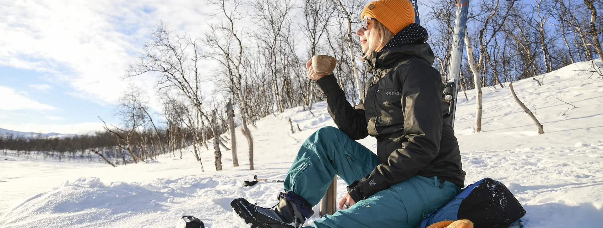 Hiihtoretkellä taukoa pitävä nainen istuu lumisessa rinteessä kuksa kädessä.
