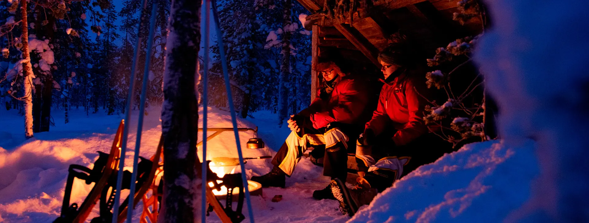 Kaksi ihmistä laavulla iltahämärässä talvisessa maisemassa.