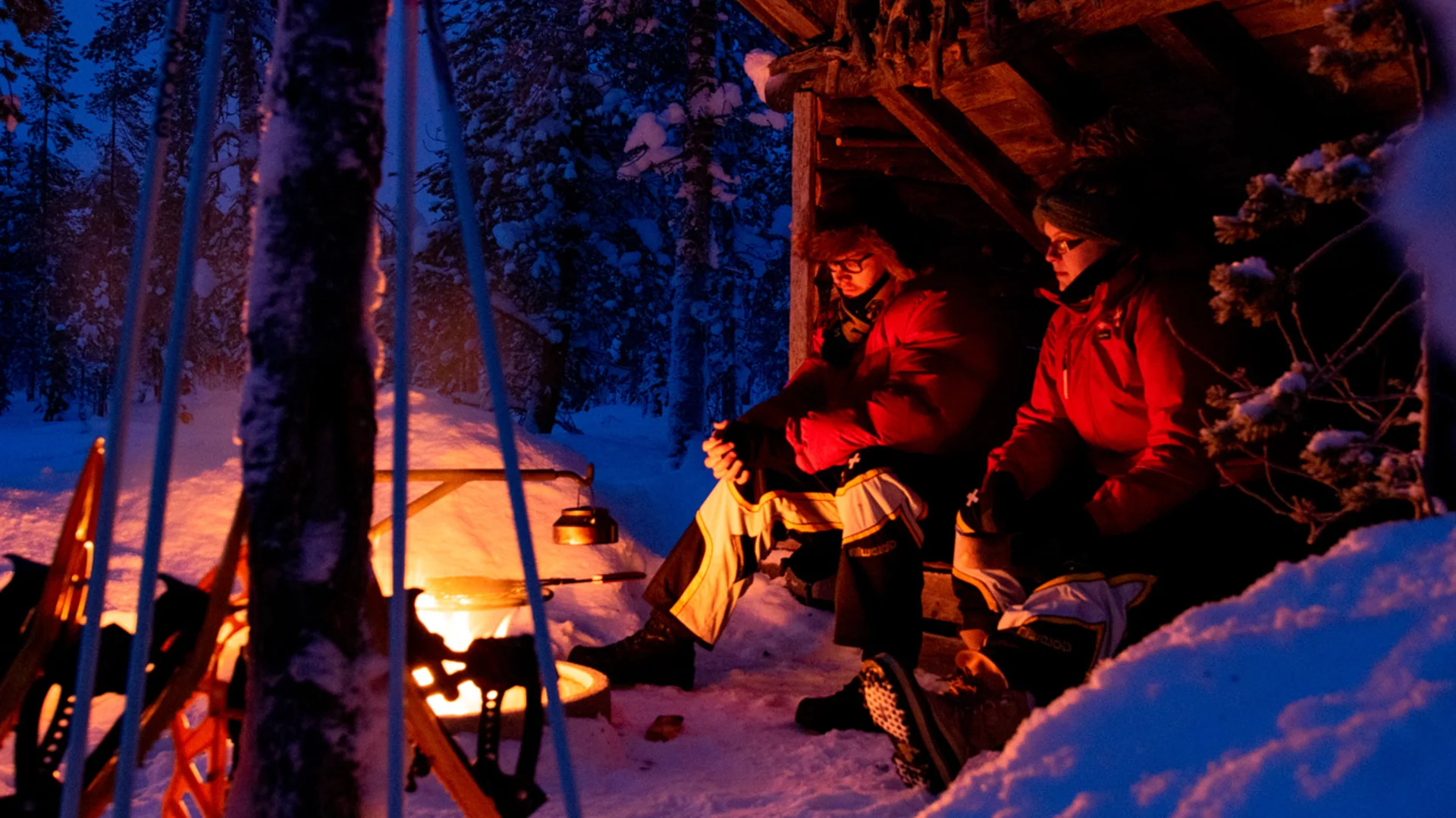 Kaksi ihmistä laavulla iltahämärässä talvisessa maisemassa.