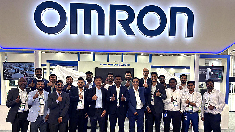 Team OMRON members smiling and giving thumbs-up in front of a large OMRON corporate logo at an exhibition booth.