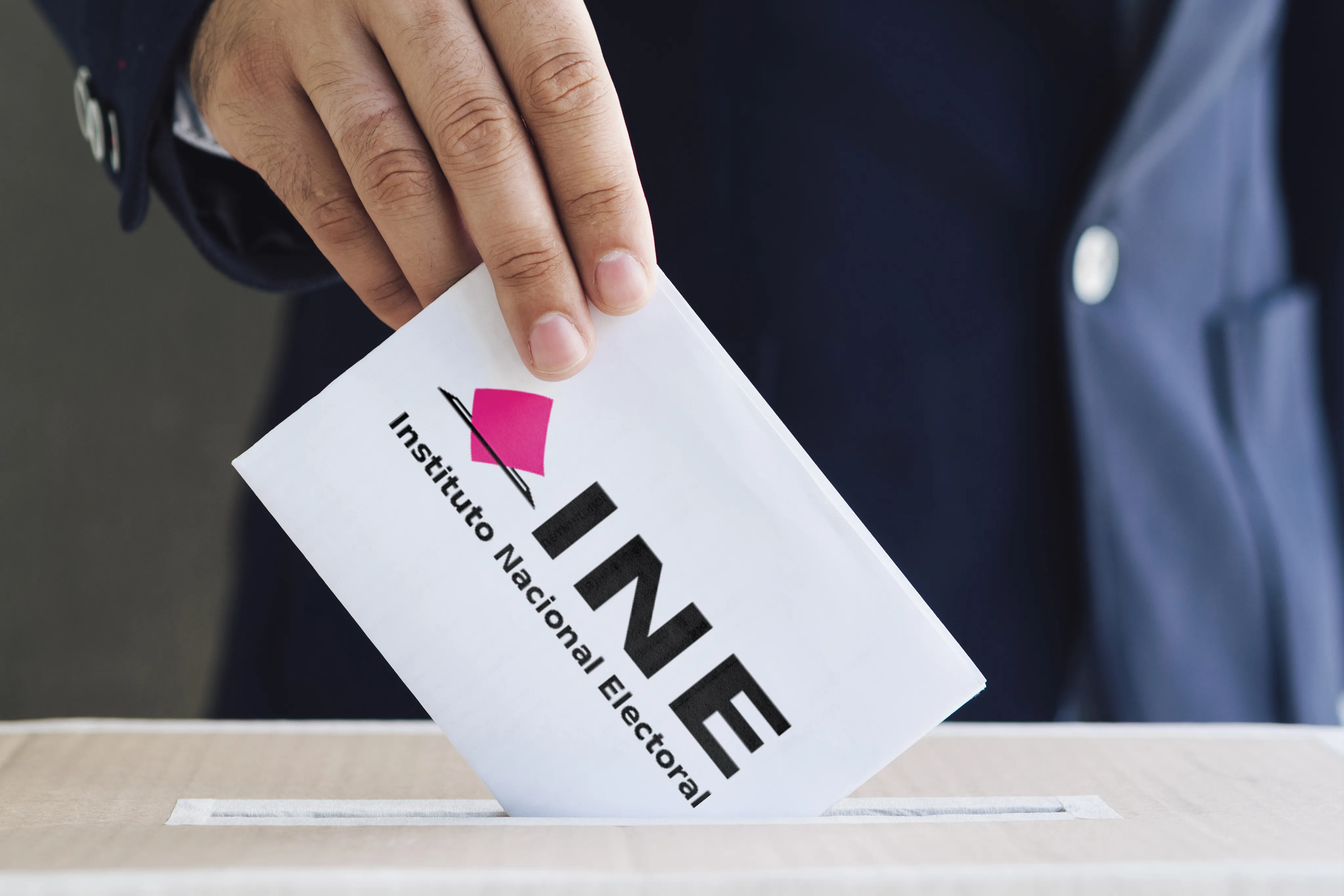 Una mano depositando un sobre con el logo del INE en una urna