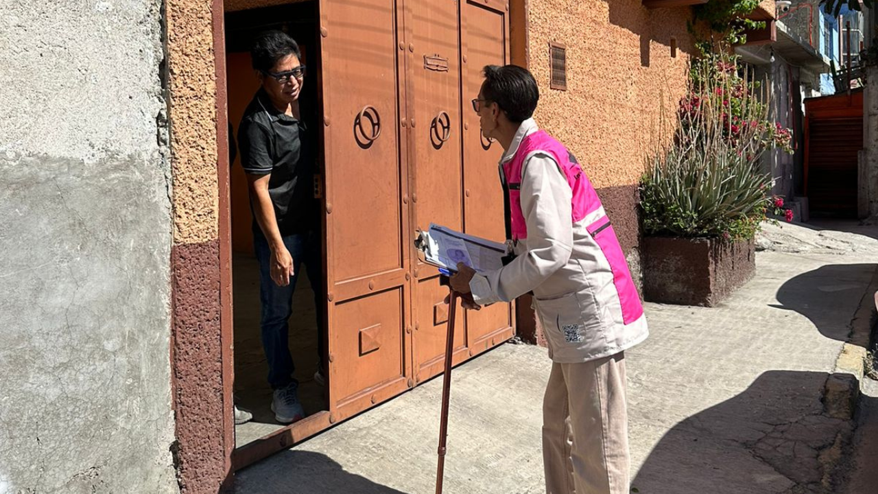 Martín (con un chaleco rosa), capacitador electoral con discapacidad, frente a un hombre recargado en la puerta de su casa.