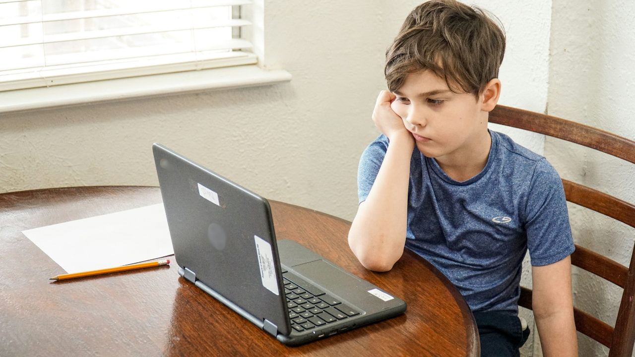 Niño sentado frente a una computadora con su cara recargada en su mano derecha en señal de aburrimiento.