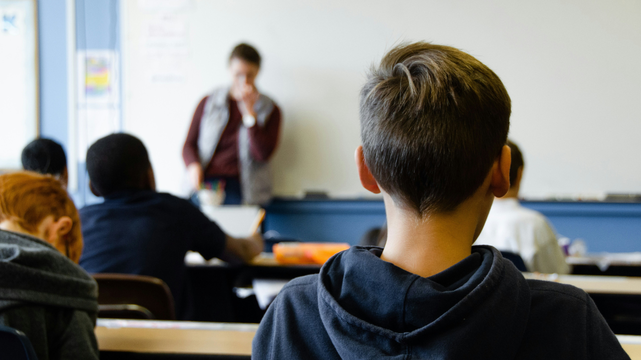 Estudiante de espaldas, frente a él, compañeros de clase y un profesor.