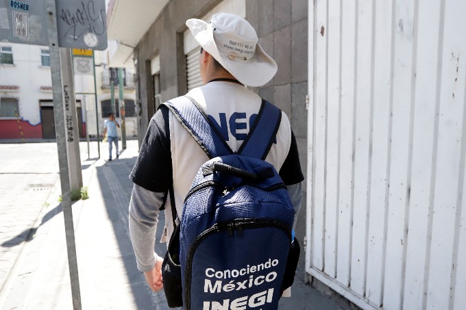 Hombre de espaldas con una mochila y chaleco en los que se lee el logo de Inegi, usando un gorro.