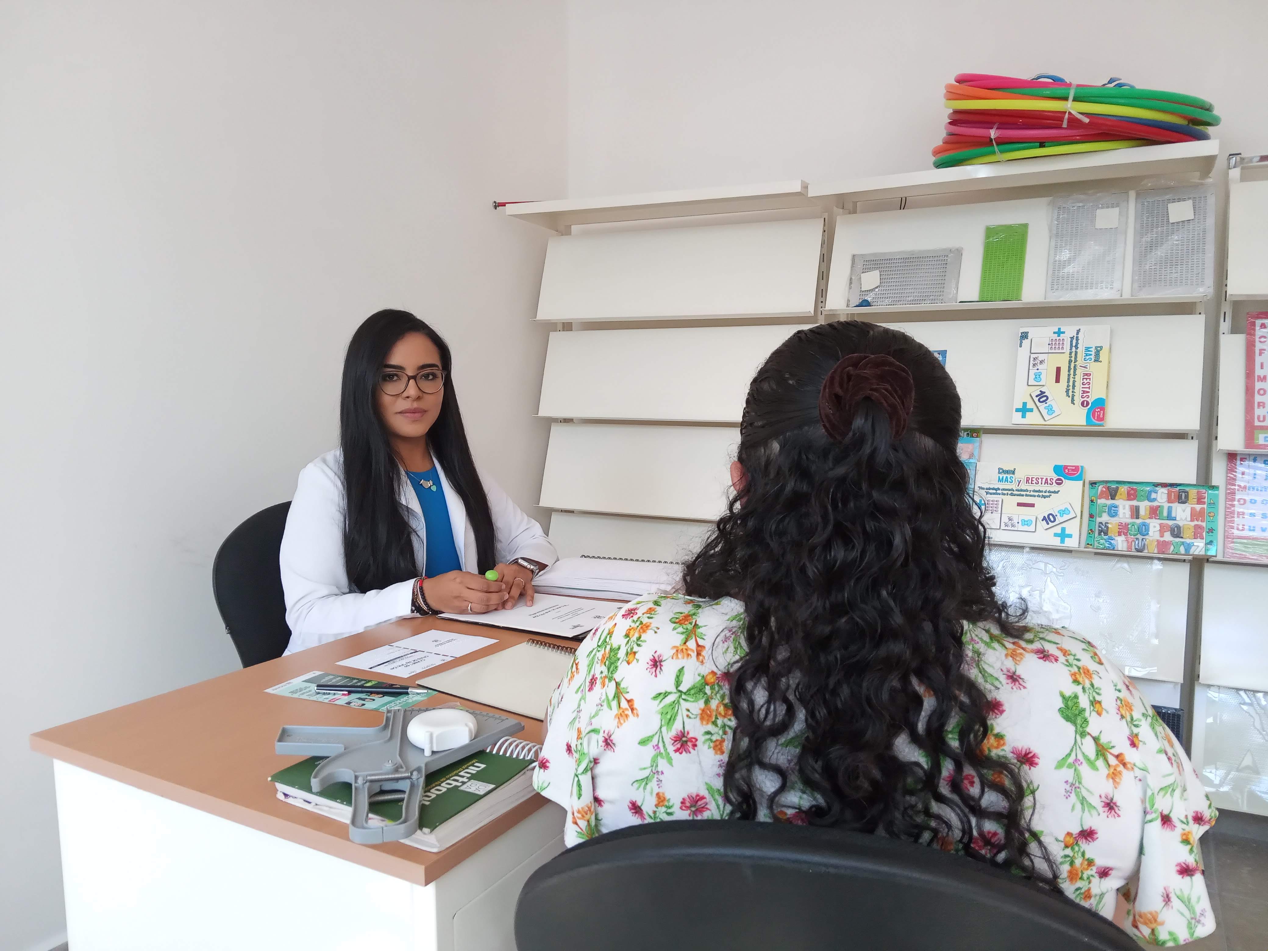 Doctora sentada de frente a una paciente mientras mira a la cámara.