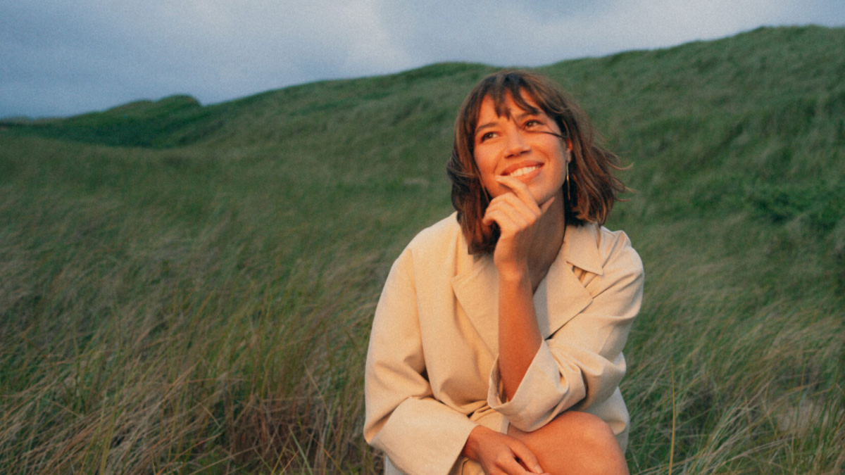 MEAU in een lichtgekleurde jas zit neer in een groen heuvellandschap.