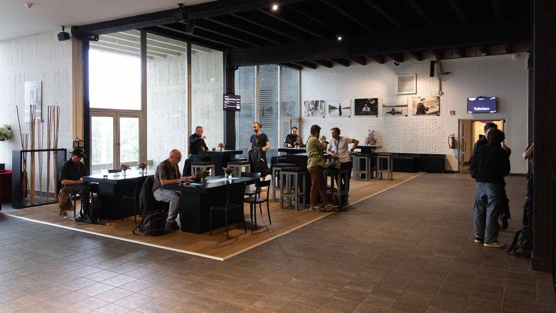 In de Verona Bar staan enkele hoge en lage tafels opgesteld. Aan de tafels staan verschillende groepjes mensen met elkaar te praten.