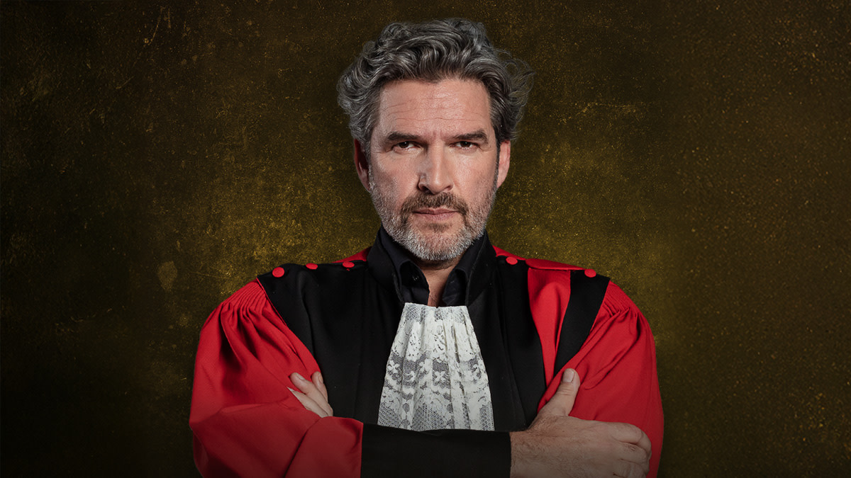 Acteur Roel Vanderstukken met een serieuze blik in een rood-zwarte advocatentoga voor de theatervoorstelling 'Assisen Live'.