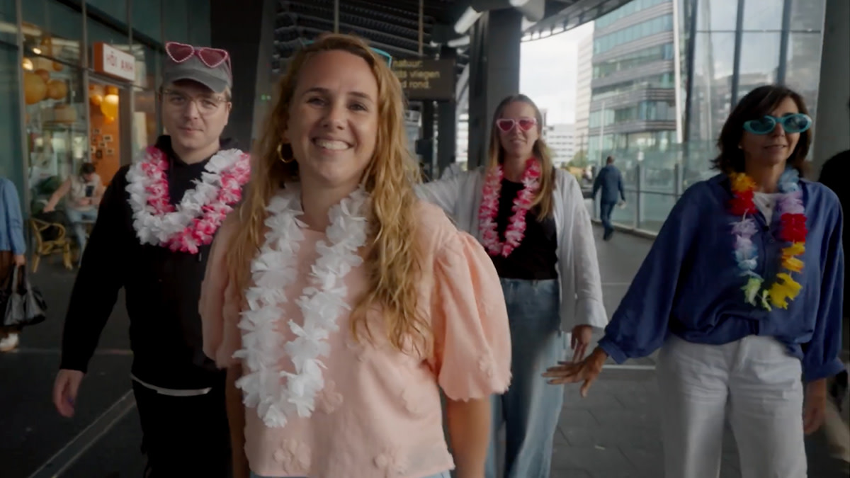 Vier collega's van be•at met een bloemenkrans rond hun hals zingen mee in een eigen videoclip bij het nummer 'Atlas' van Pommelien Thijs