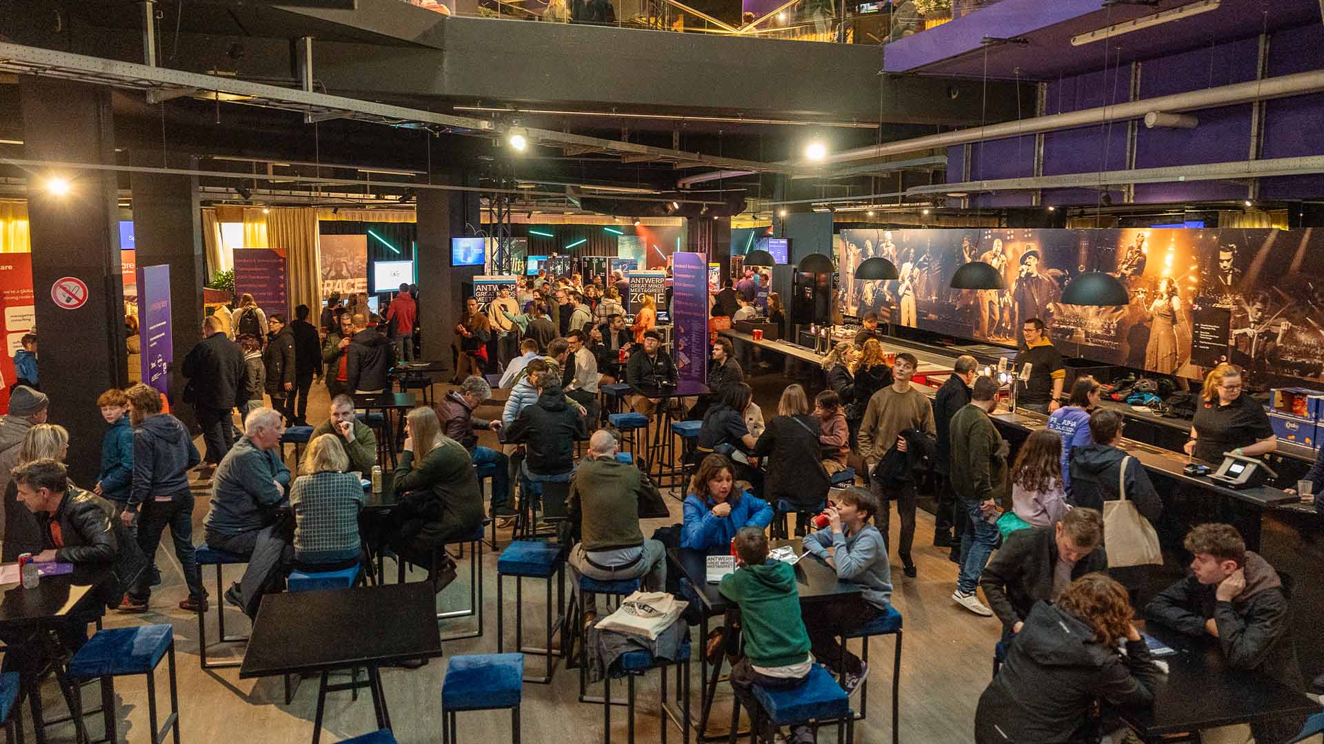 A wide view of the bustling Hospitality Center with many visitors sitting at tables, standing, and interacting, surrounded by displays and lighting.
