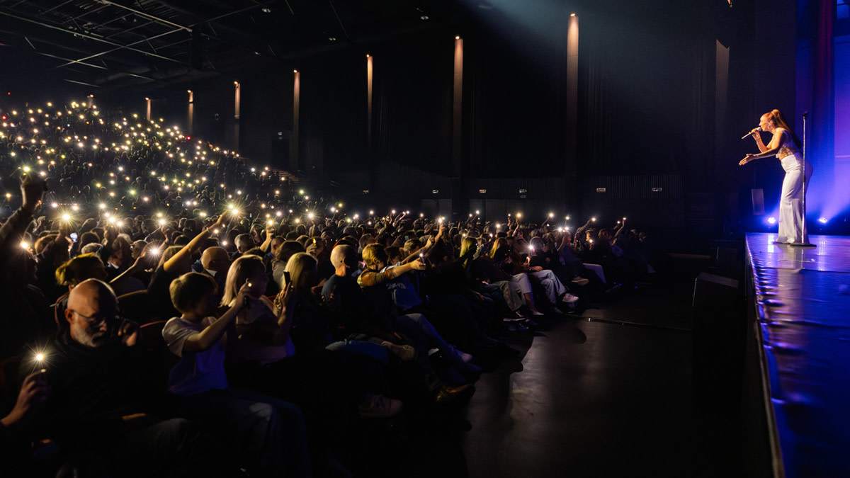 Zangeres op podium, verlicht door blauwe spotlight, tijdens een donker concert. Publiek houdt massaal smartphones omhoog met de flitslichten aan, wat een schitterend lichteffect creëert in de zaal.