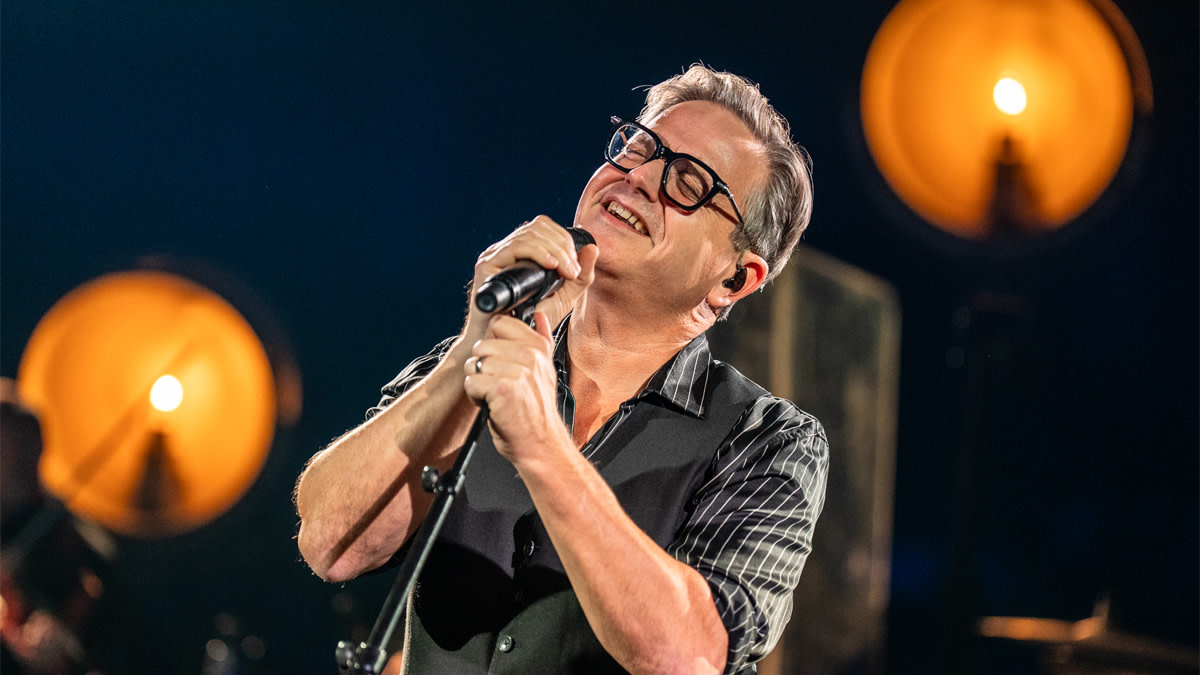 Guus Meeuwis tijdens een intieme mini-showcase in AFAS Dome, glimlachend terwijl hij in de microfoon zingt.