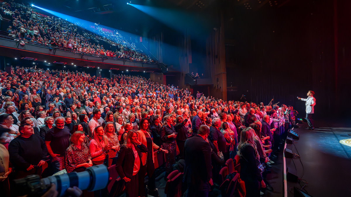 Overzichtsfoto van een live optreden in een theaterzaal. Een artiest staat op het podium en groet een uitverkocht, enthousiast publiek. De zaal is gevuld met toeschouwers die staan en zitten, met felle rode en blauwe podiumverlichting.