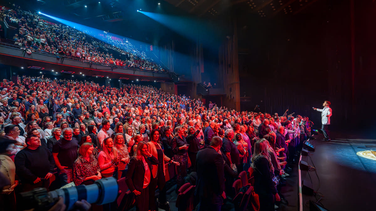 A standing ovation from a sold-out audience at Stadsschouwburg Antwerp during a live performance, with the artist on stage and the audience visible all the way up to the balconies.