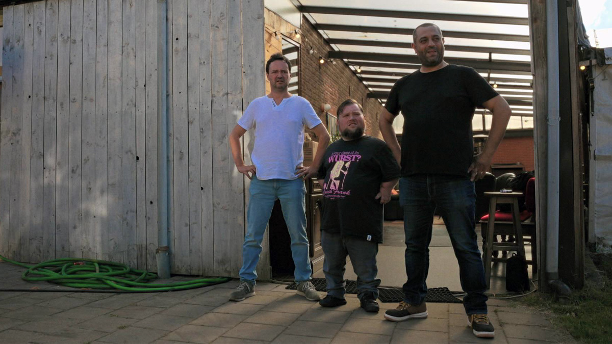 Han Solo, William Boeva en Erhan Demirci staan naast elkaar voor een backstagegebouw tijdens de opnames van de Streamz-reeks Onzen Tour. De drie comedians poseren ontspannen in openlucht, met achter hen een terras en kleedruimte.