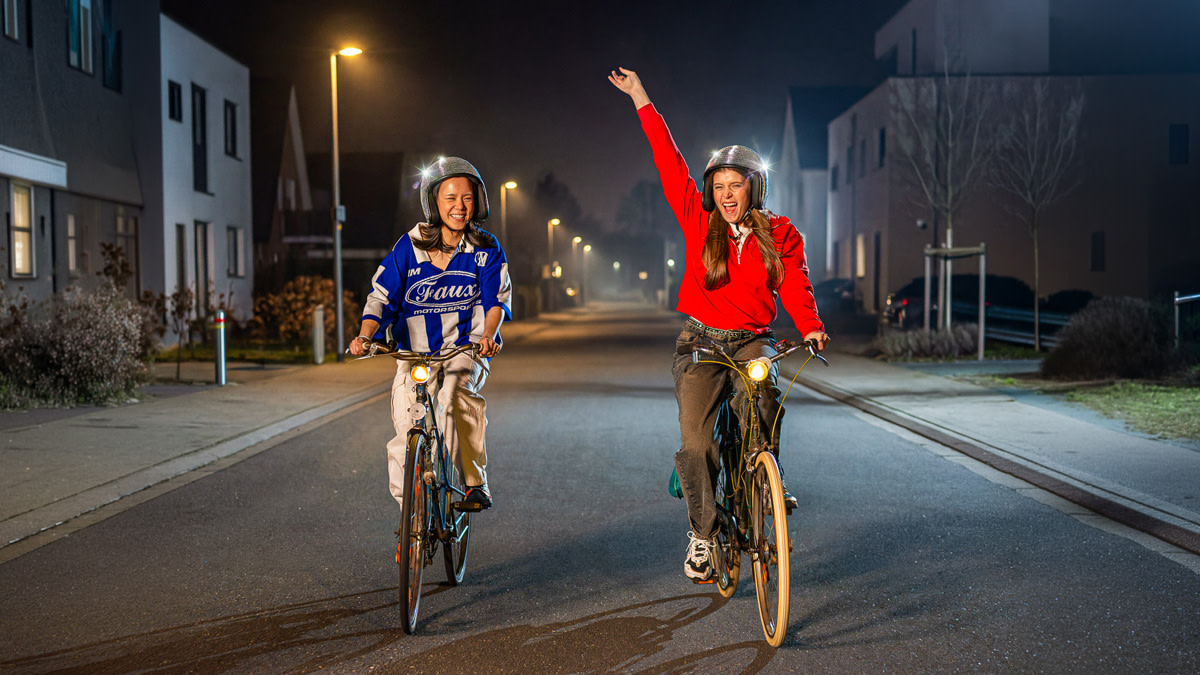 Two cheerful women are cycling down a quiet street in the evening, with their bike lights on and clearly enjoying themselves. One of them enthusiastically raises her arm in the air as they seem to be cycling together towards an evening full of excitement.