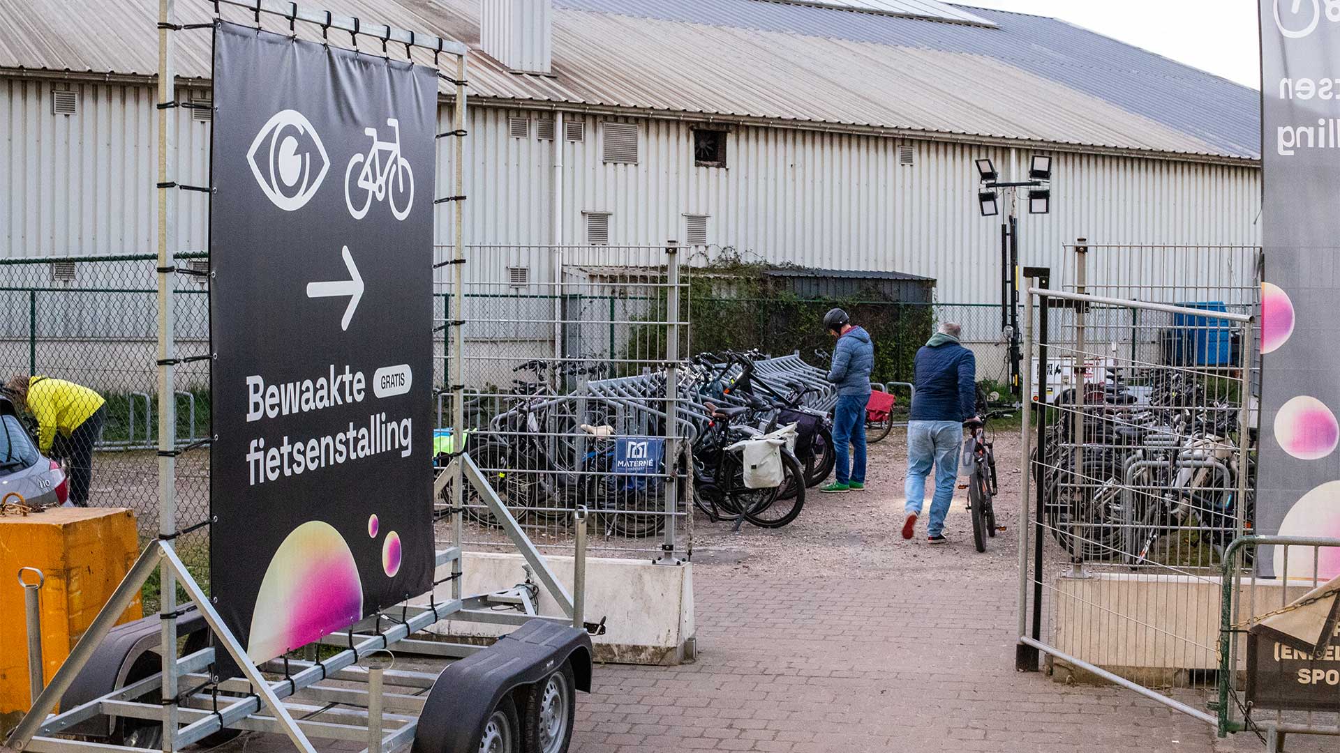 Entrée du parking vélos surveillé de l’AFAS Dome sur le parking Vaart, avec un panneau directionnel indiquant « Bewaakte fietsenstalling », des vélos dans des supports et des visiteurs qui stationnent leur vélo.