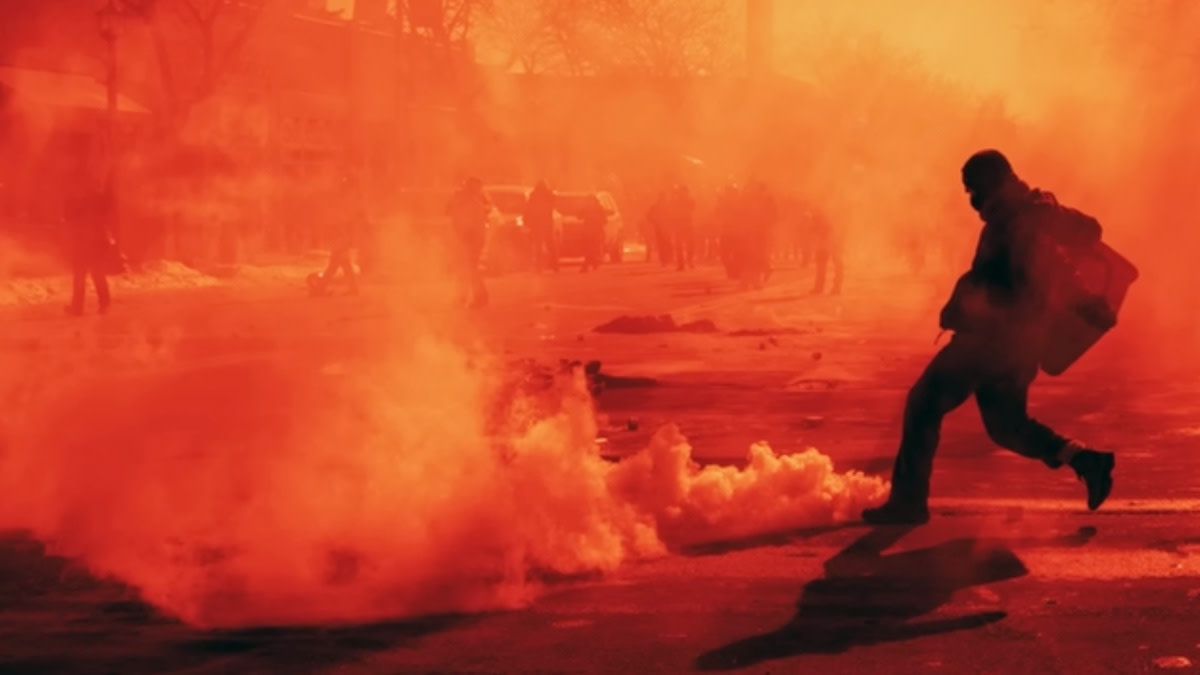 Een demonstrant loopt door een straat vol rook en traangas terwijl op de achtergrond protestscènes zichtbaar zijn in een rood-oranje gloed.