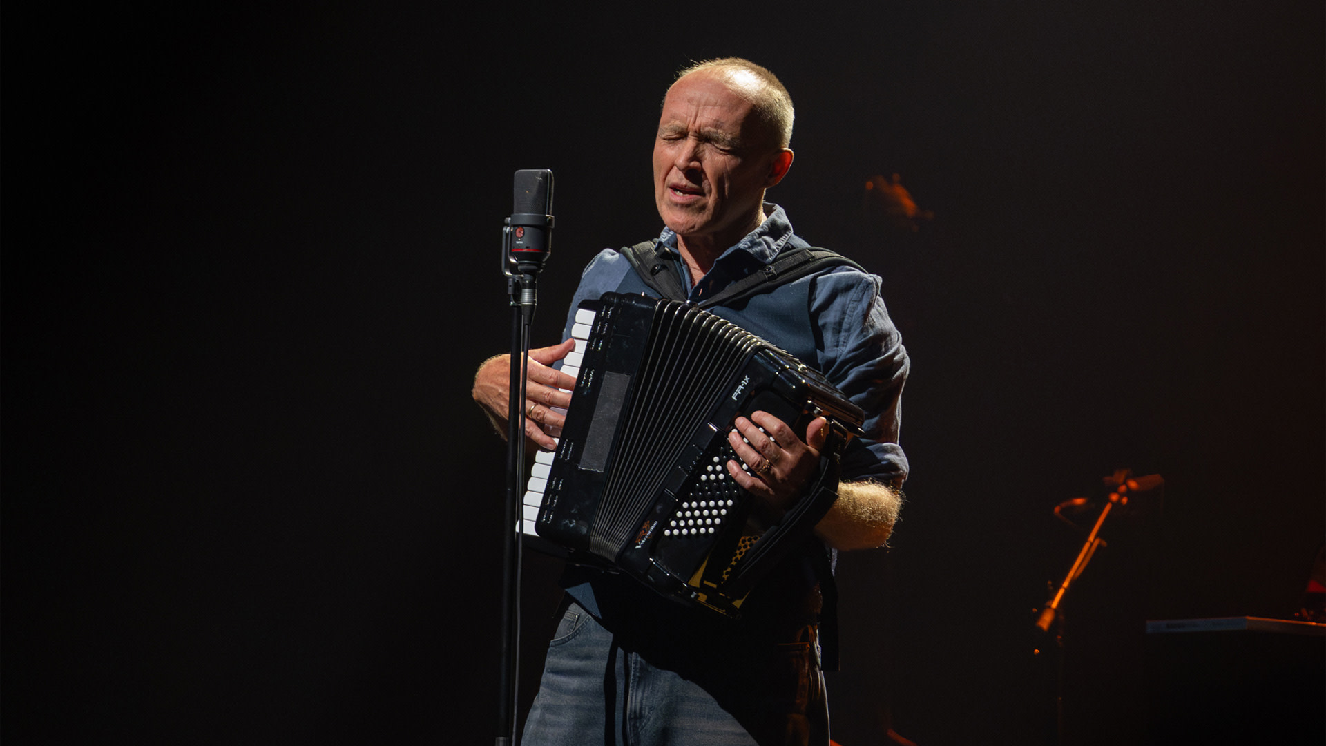 Stef Bos zingt en speelt accordeon tijdens een liveconcert op een sfeervol verlicht podium.