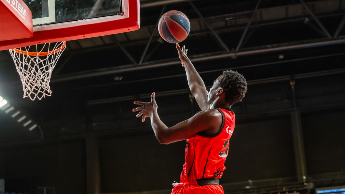 Een basketballer van Antwerp Giants in een rood tenue zweeft richting de basket voor een lay-up in een donkere Lotto Arena, met de bal vlak voor de ring tijdens een intense wedstrijd.