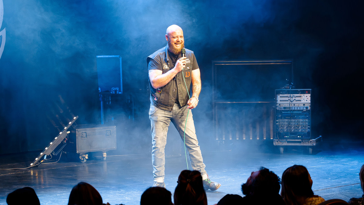 Comedian Lev Vanorbeek in zijn biker-outfit met een microfoon in de hand op een theaterpodium.