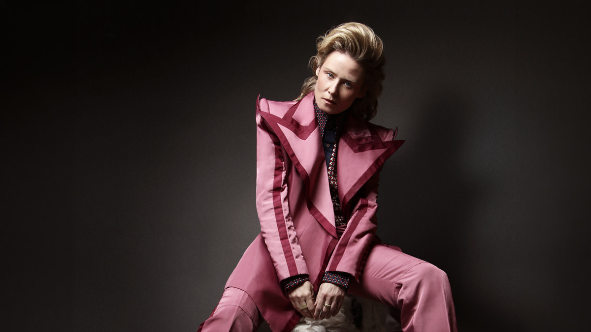 Róisín Murphy zit in een licht paars rozig pak op een barkruk tegen een zwarte achtergrond.