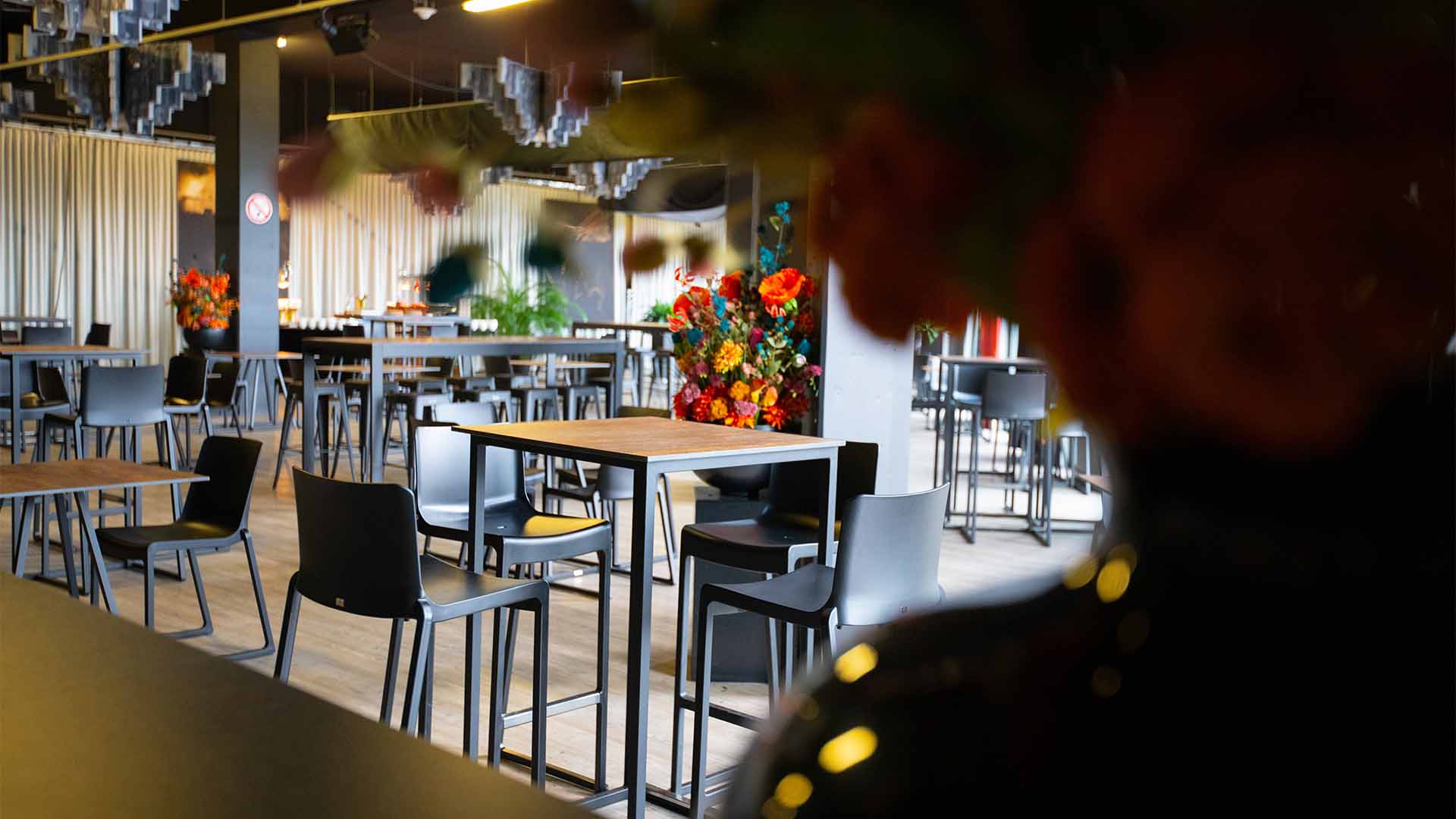 Een sfeervol ingerichte evenementenruimte in de Lotto Suites, met moderne hoge tafels en stoelen, en kleurrijke bloemstukken als decoratie.