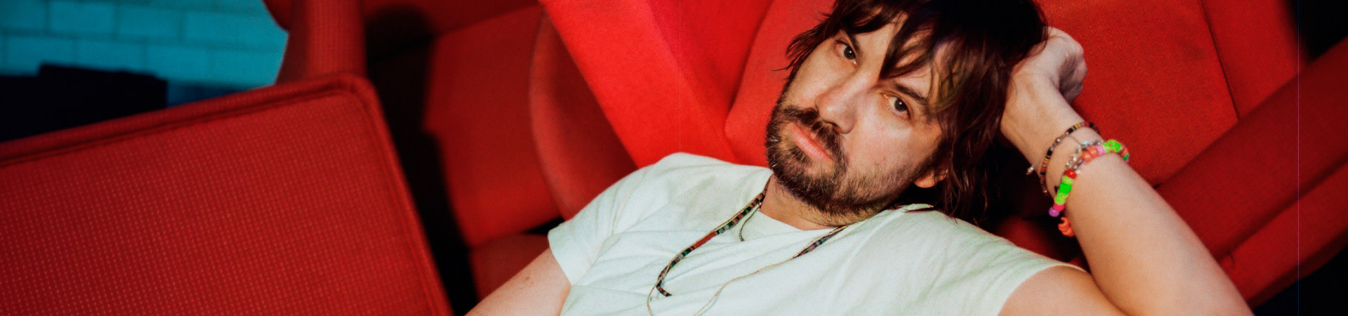 Homme barbu allongé dans un fauteuil rouge, regardant la caméra, portant des bracelets colorés dans une ambiance artistique et intime.