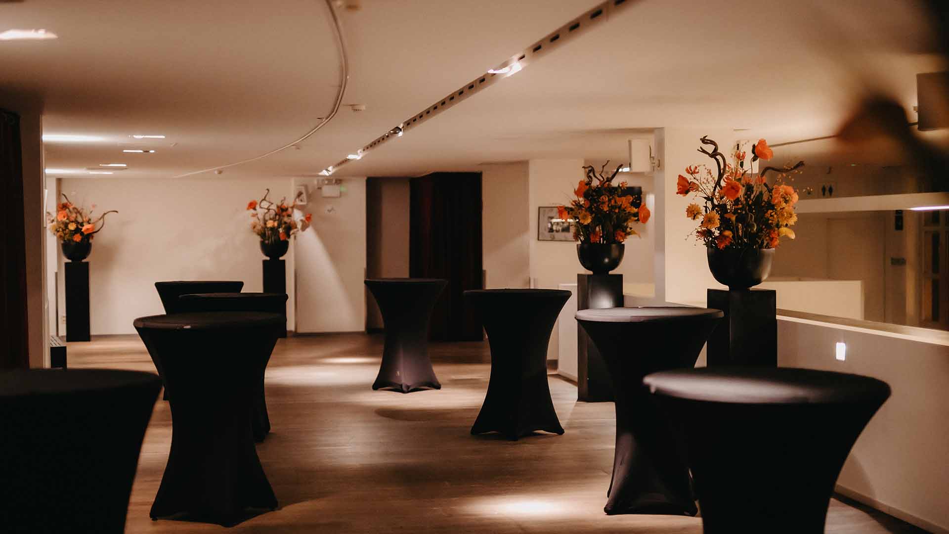 Stijlvol ingerichte foyer in Capitole Gent met hoge statafels en bloemstukken in zwarte vazen op sokkels, onder warme sfeerverlichting.