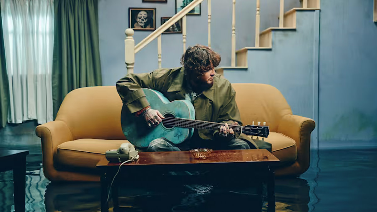 James Arthur zit met zijn gitaar in een lichtbruine zetel in het midden van een woonkamer die onder water staat.
