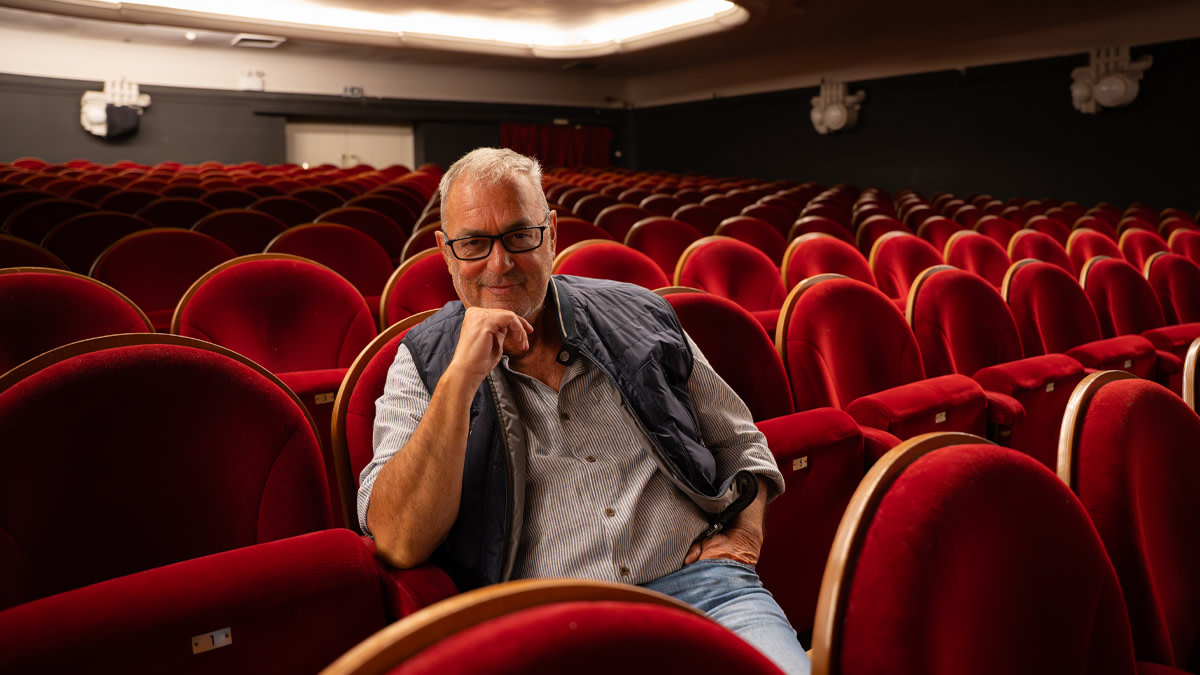 Zanger Bart Herman zit glimlachend tussen de rode theaterstoelen, met zijn hand tegen zijn gezicht leunend, in een lege theaterzaal.