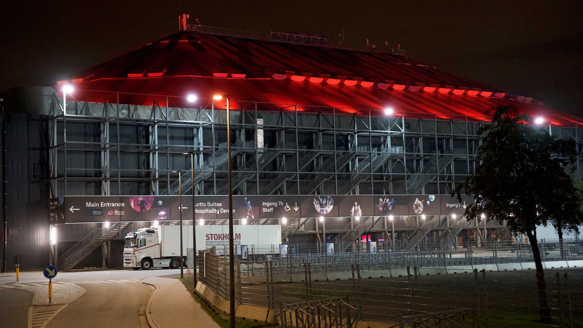 AFAS Dome bij nacht met een dak dat fel oranje verlicht is. Het gebouw toont metalen constructies en trappen, met enkele verlichte borden en een vrachtwagen geparkeerd voor de ingang.