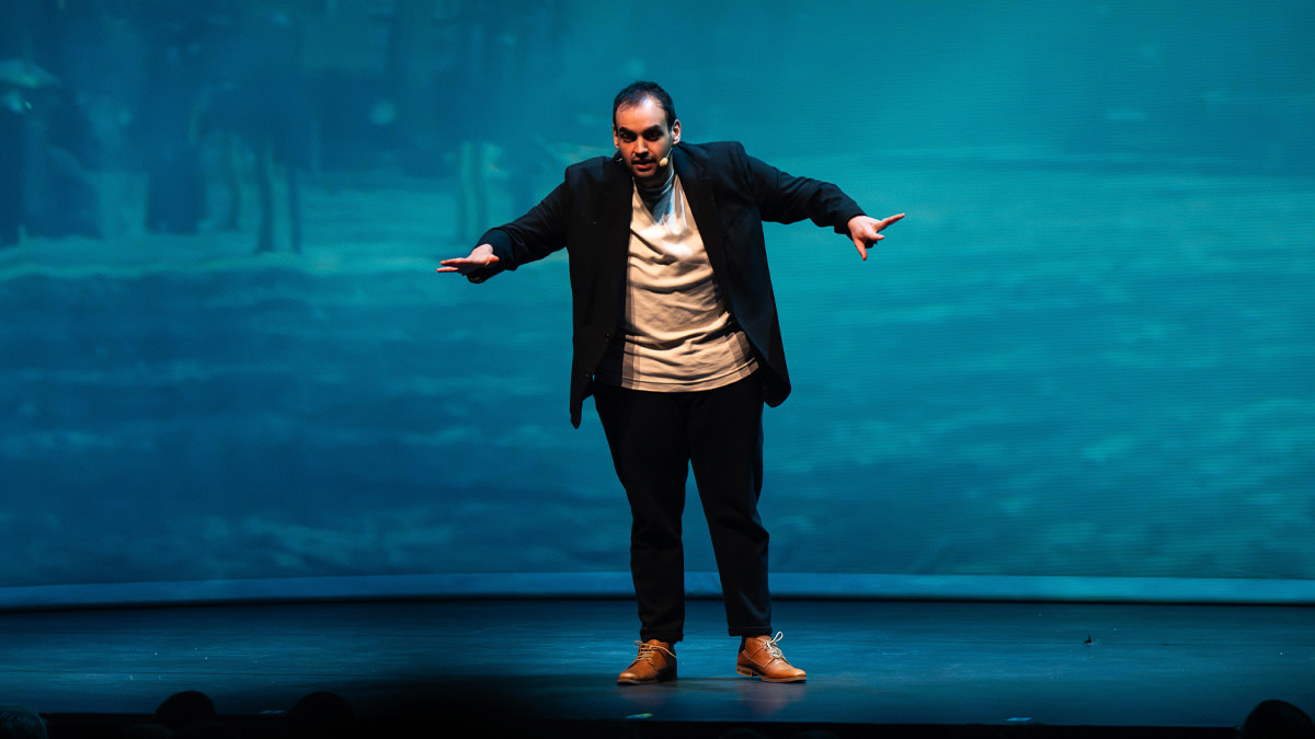 Kamal Karmach staat in zwart pak tegen een blauwe achtergrond op het podium in Capitole Gent.