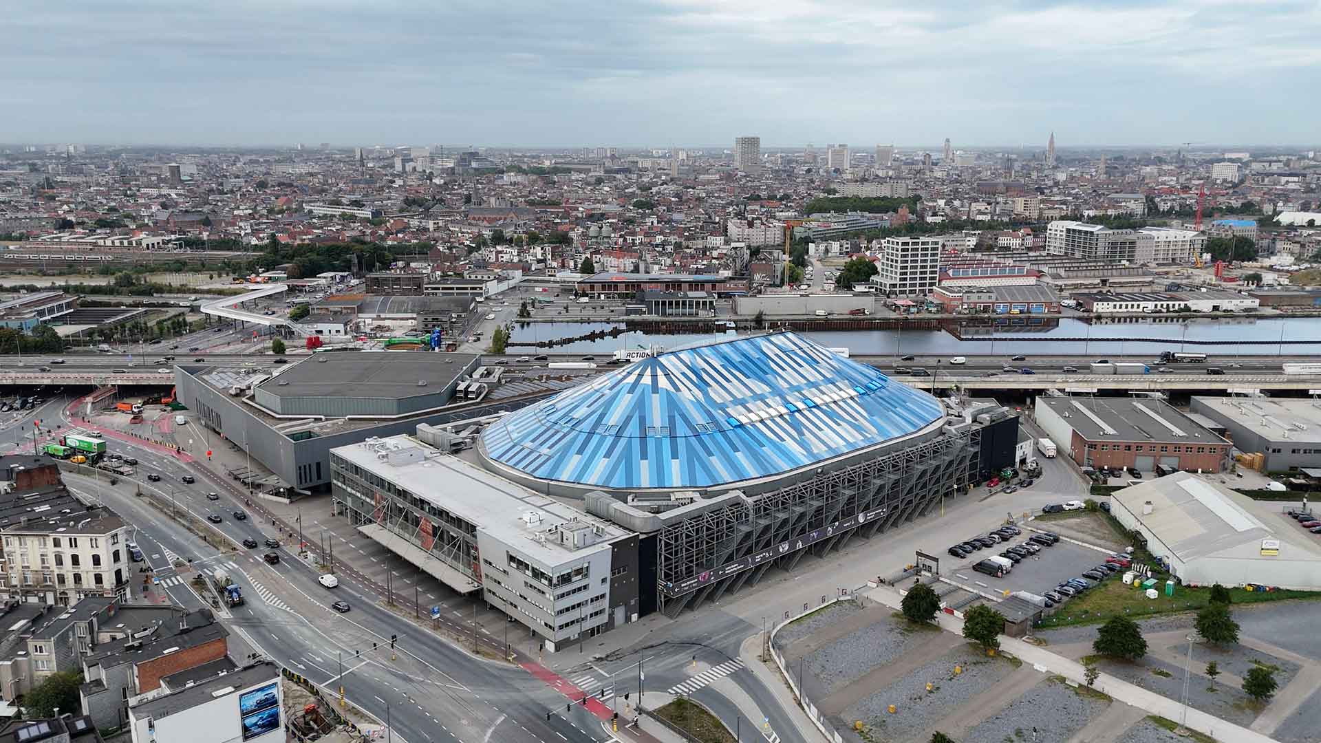 De AFAS Dome, een grote concertzaal met een opvallend blauw en wit gestreept dak, is te zien in een stedelijk landschap. De foto is genomen vanaf een verhoogd standpunt en toont een stad met veel gebouwen en wegen.