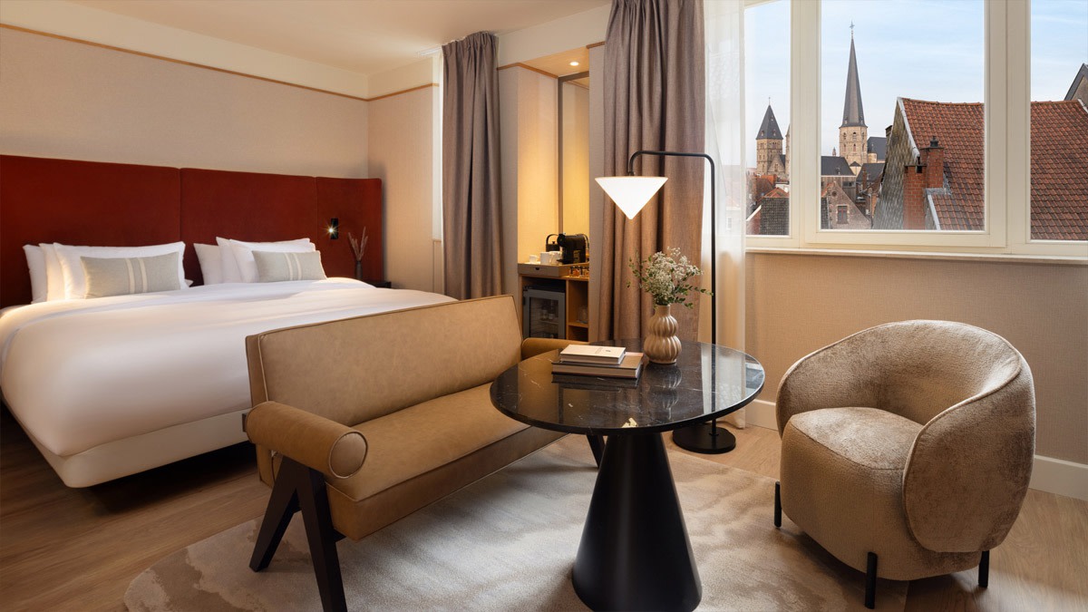 Chambre élégante au NH Collection Gent avec grand lit, coin salon avec fauteuil et table, éclairage chaleureux et vue sur toits historiques et clochers.